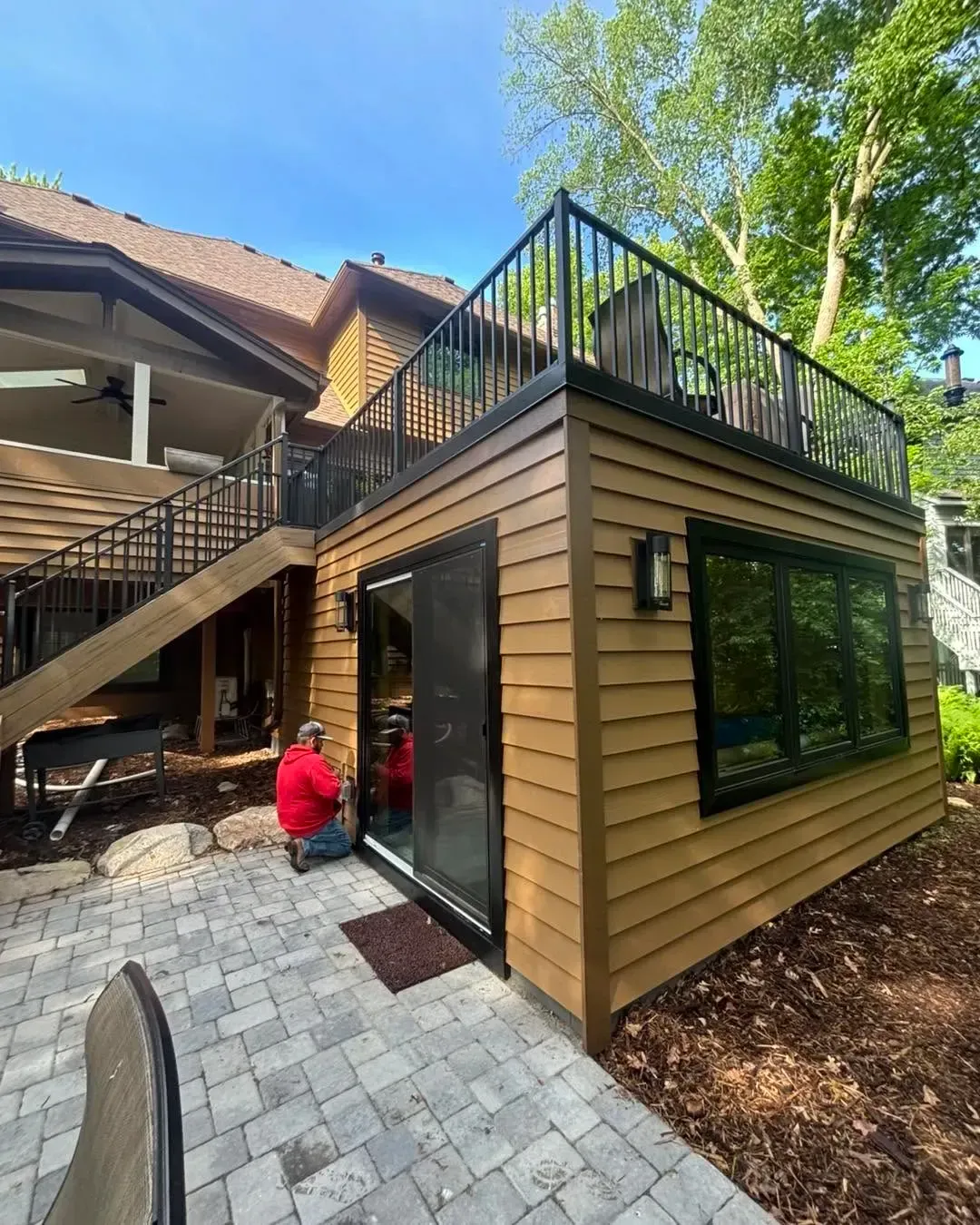 Modern tan-sided patio addition with rooftop deck, black railings, and large windows beside brick pavers